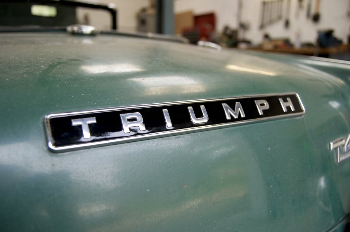 Black TRIUMPH badge with chrome letters and surround, fitted to the green bonnet of a classic car