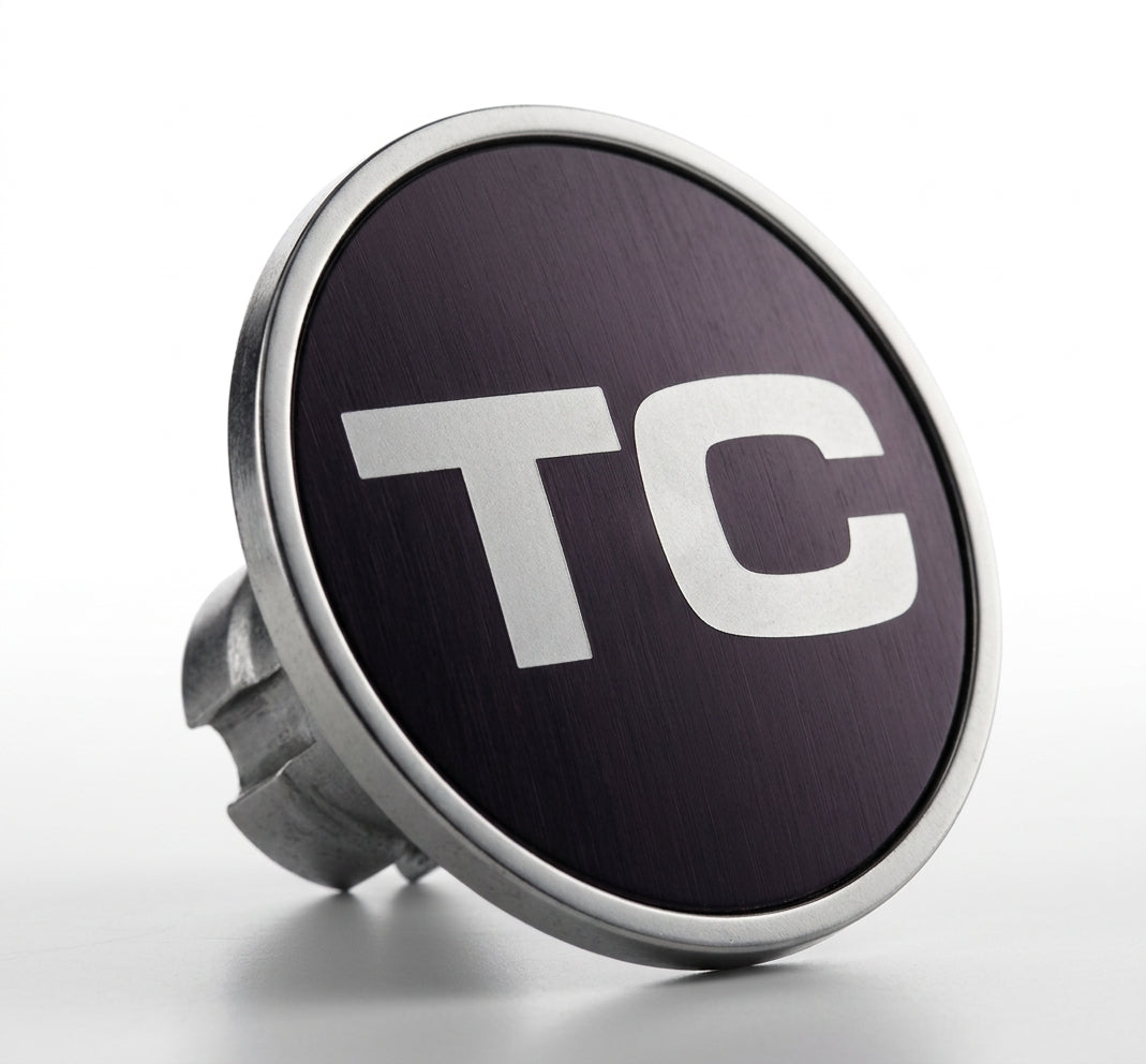 Round chrome-rimmed badge reading TC, angled studio shot on white background