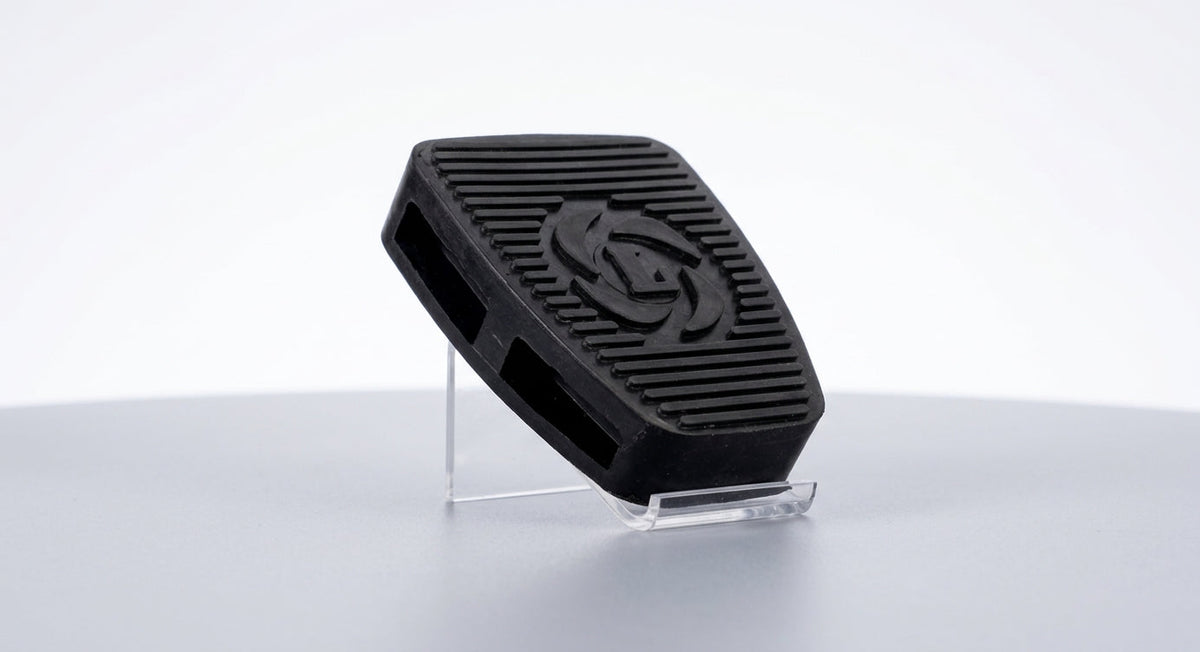 Black ribbed rubber pedal pad with circular logo motif, studio shot on clear stand