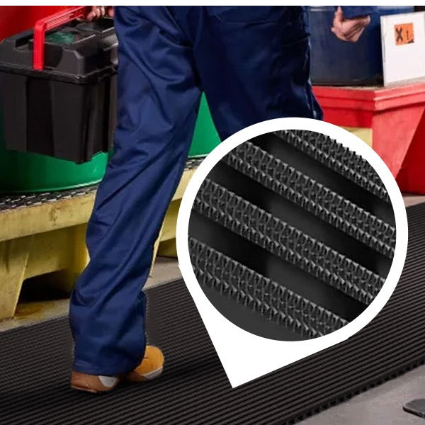 A worker in blue overalls and work boots is walking and carrying a toolbox over a black, heavy-duty industrial matting roll in a factory or warehouse. The mat provides non-slip, high-traction footing, which is essential near chemical storage. An inset circle magnifies the mat's UV-resistant surface with diamond-patterned ribs, confirming its suitability for wet areas, industrial, and outdoor use.