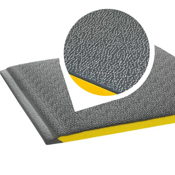 Close-up, angled view of the corner of a heavy-duty anti-fatigue mat showing the dark grey, pebbled, non-slip PVC foam surface and the bright yellow foam base layer. An inset magnifies the wear-resistant texture and thick construction, emphasizing cushioned support for workstations, garages, and industrial use.