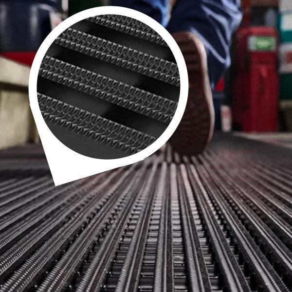Close-up of a black, heavy-duty industrial matting roll laid out on a workshop floor next to a bright orange tool chest. Two workers are blurred in the background. An inset circle magnifies the mat's non-slip, high-traction surface, featuring UV-resistant diamond-patterned ribs, ideal for wet areas and industrial or outdoor use.