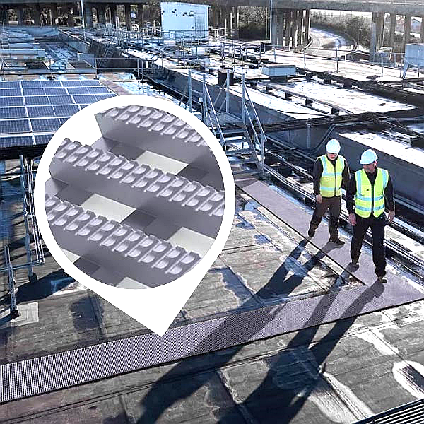 Grey anti-slip walkway matting on an industrial rooftop. Two workers in safety vests walk on the non-slip fiberglass planking next to solar panels, providing a secure path on the commercial roof near a bridge structure.
