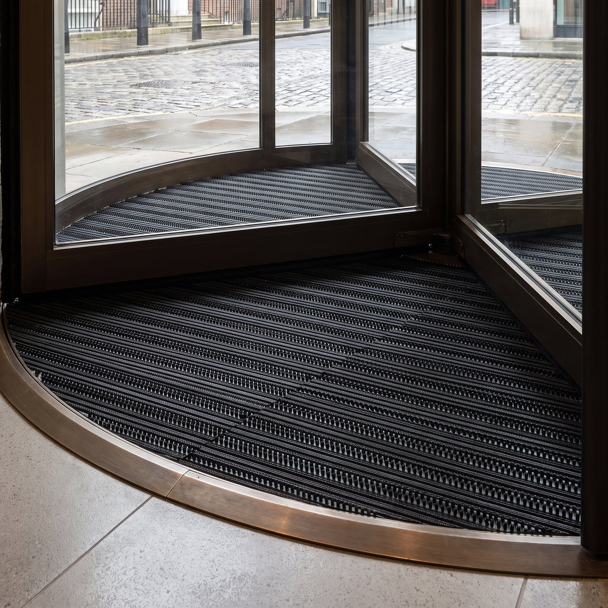 Black brush insert entrance matting, standard grid, recessed in a curved matwell at the foot of a commercial revolving door