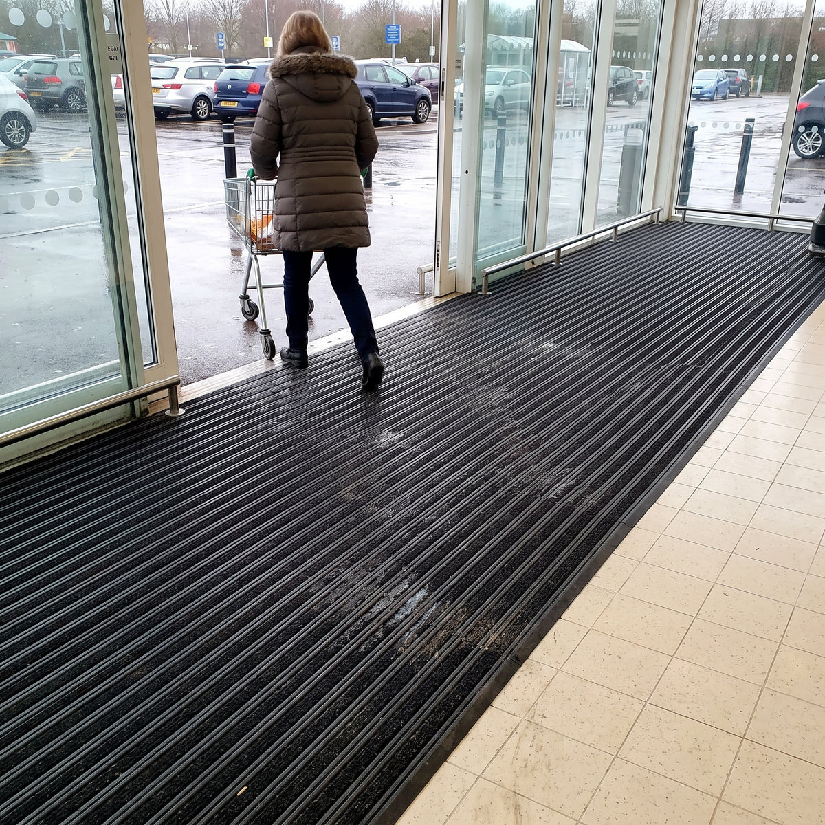 Black carpet insert entrance matting laid as a long run inside a supermarket entrance, shopper pushing a trolley in from a wet car park