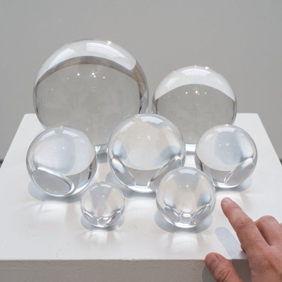 Seven clear acrylic spheres in graduated sizes grouped on a white plinth, a hand reaching in at the right edge