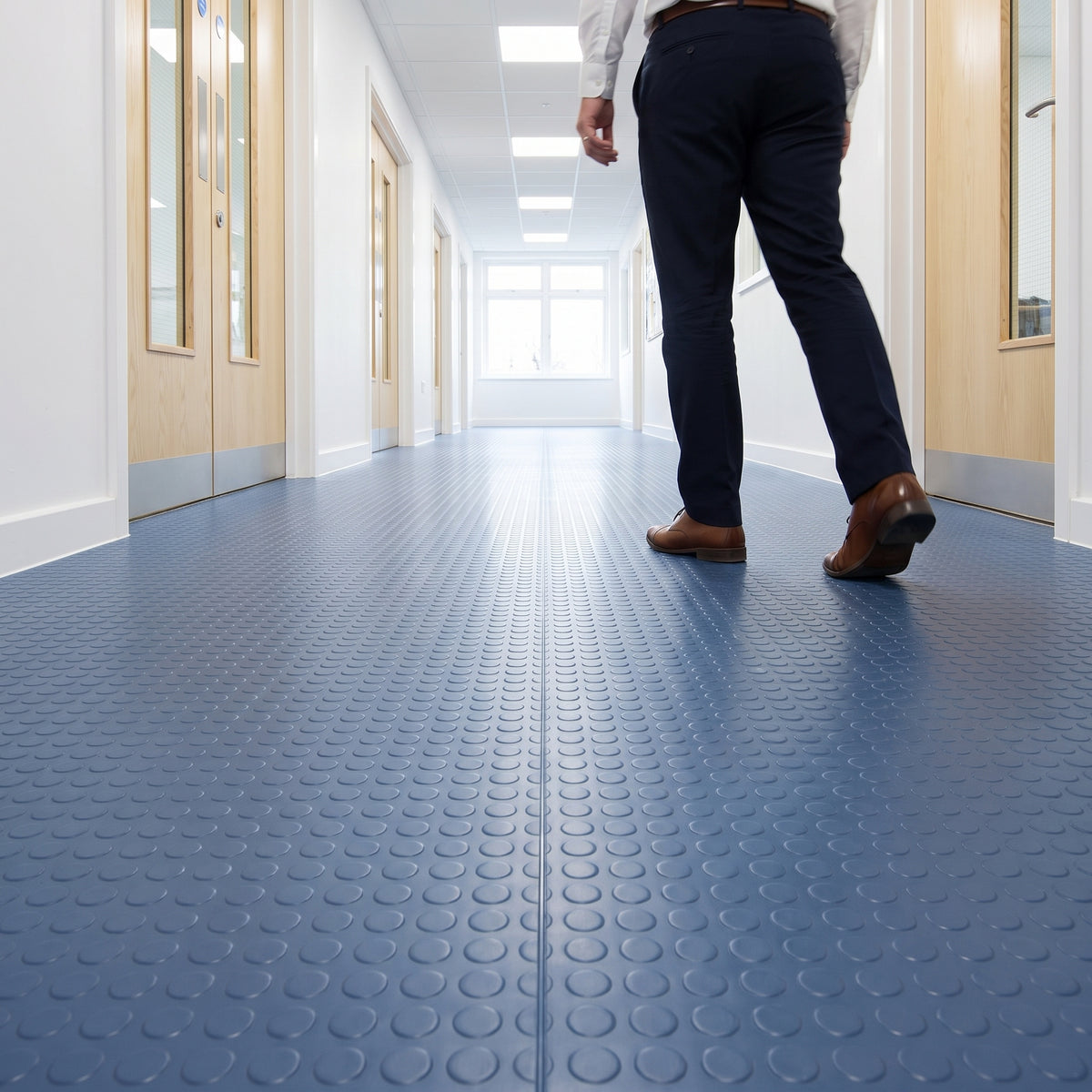 Blue coin-pattern PVC floor matting laid in wide roll lengths down a bright school corridor with oak doors