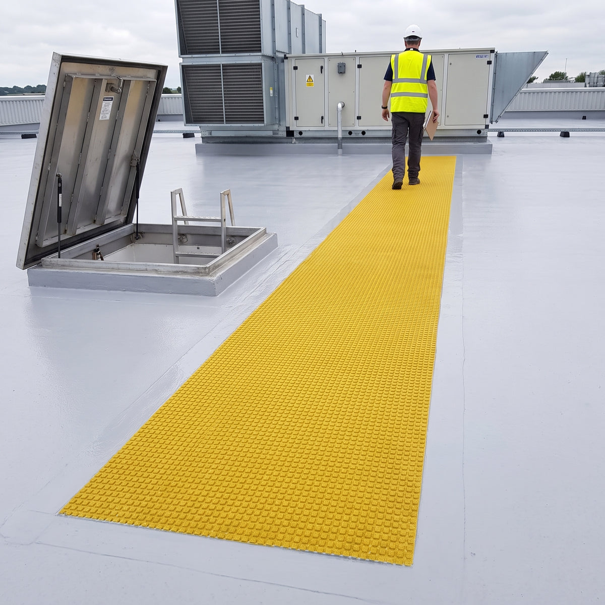 Roof inspector in hi-vis and safety helmet walking a single straight run of yellow embedded walkway matting bedded flush into a pale liquid-applied roof coating, from the roof hatch to an air-handling unit