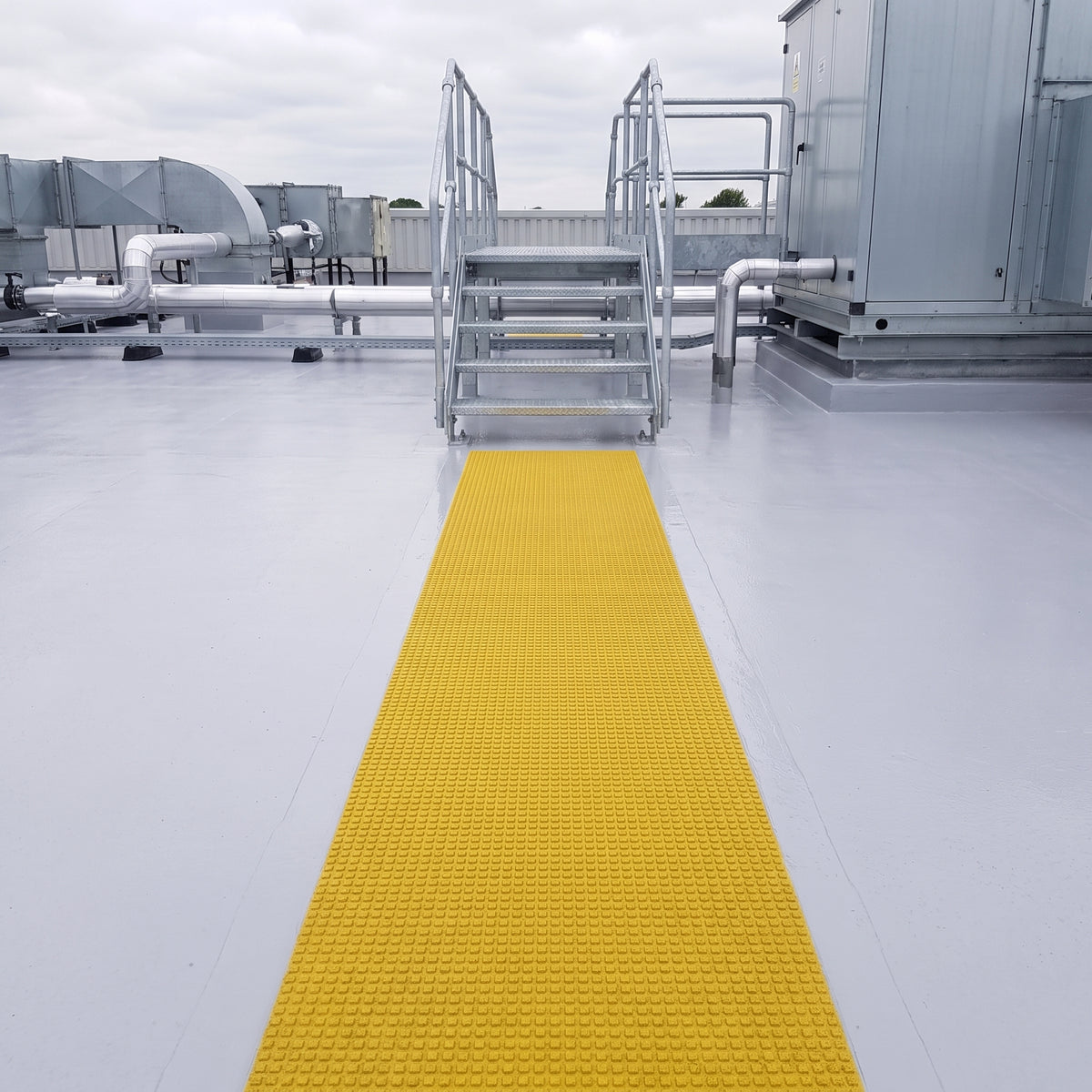 Yellow embedded walkway matting running straight across a pale flat roof to the foot of a galvanised plant access stair
