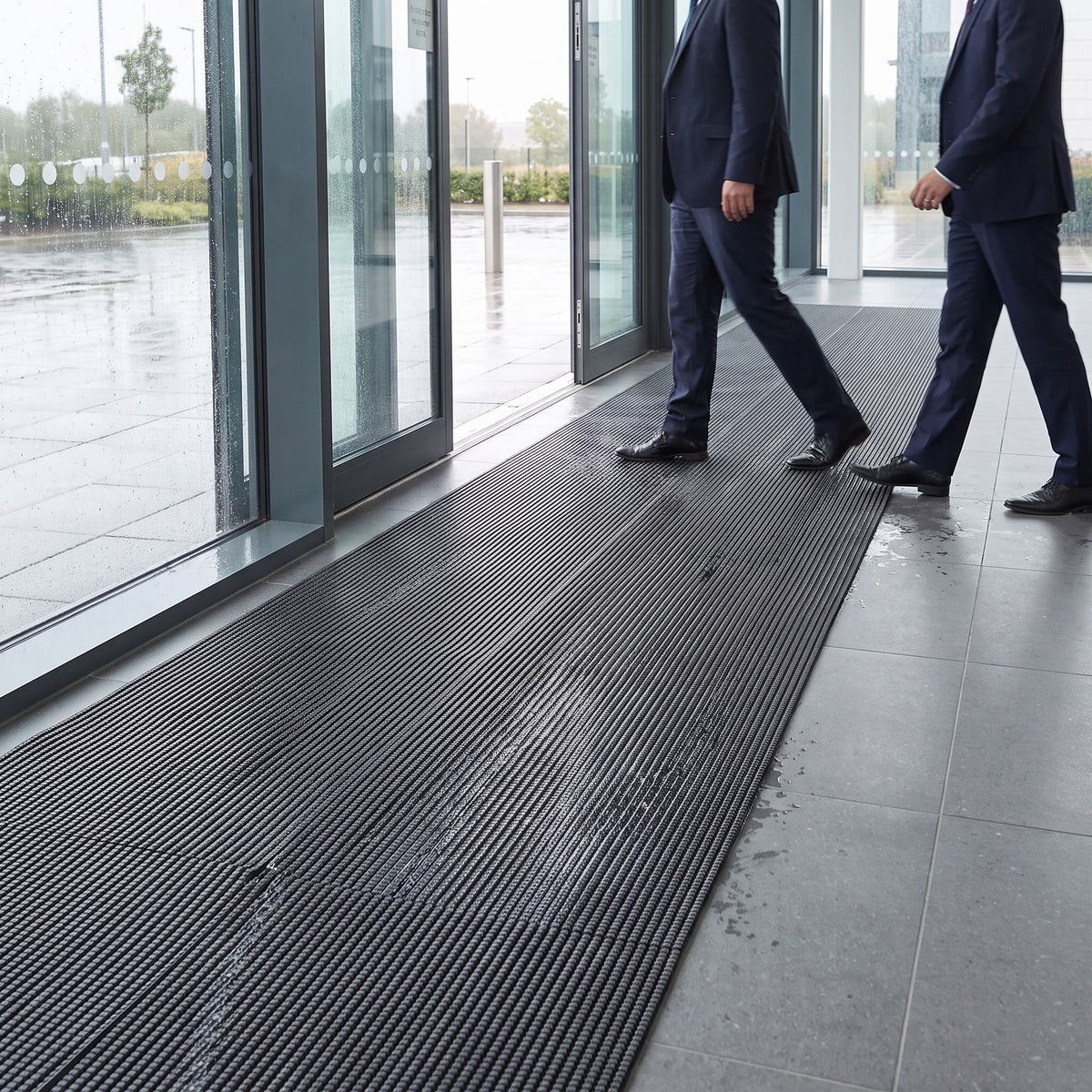 Dark grey open-grid entrance matting laid across a glazed office lobby, two visitors walking in from the rain