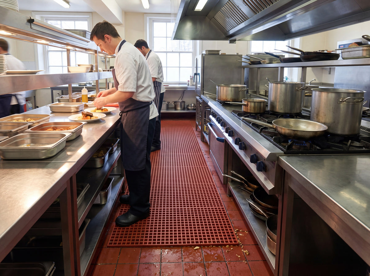 Red open grid PVC matting laid along a commercial kitchen hot line, food debris falling through to the tiled floor