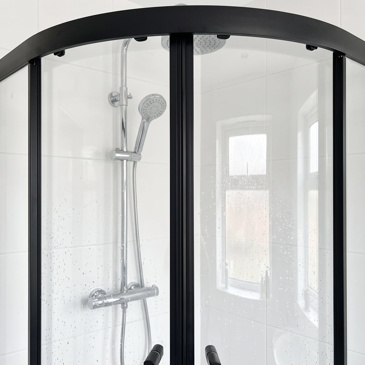 Closed black-framed quadrant shower enclosure with curved head rail, chrome riser rail and window behind
