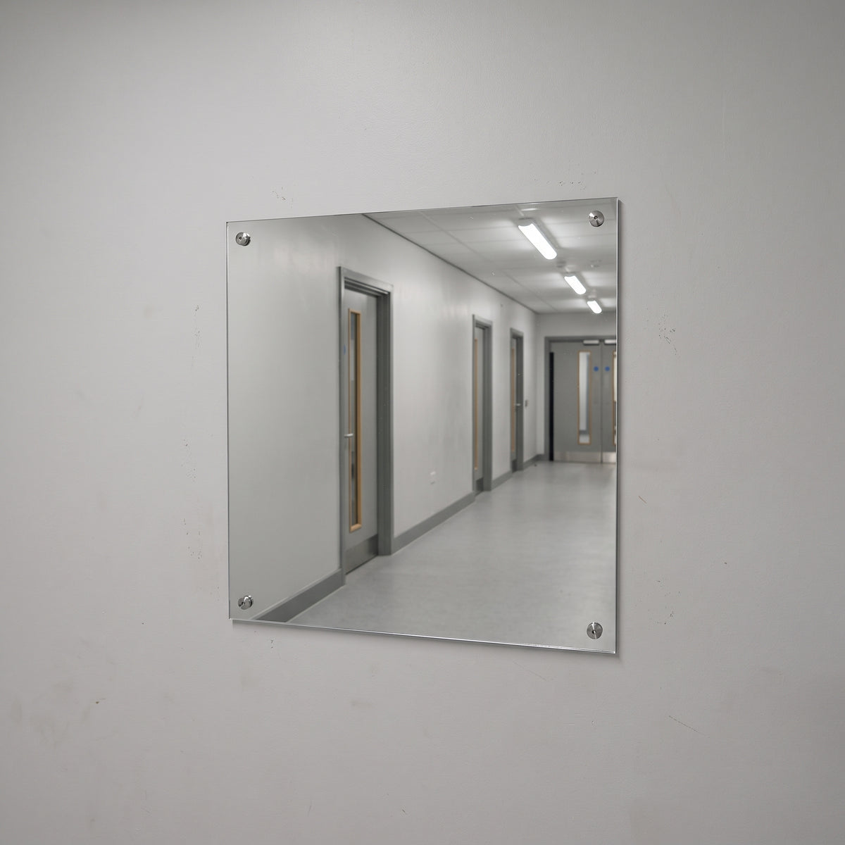 Silver polycarbonate mirror screw-fixed to a plain wall, reflecting a lit corridor with pale timber doors and strip lights