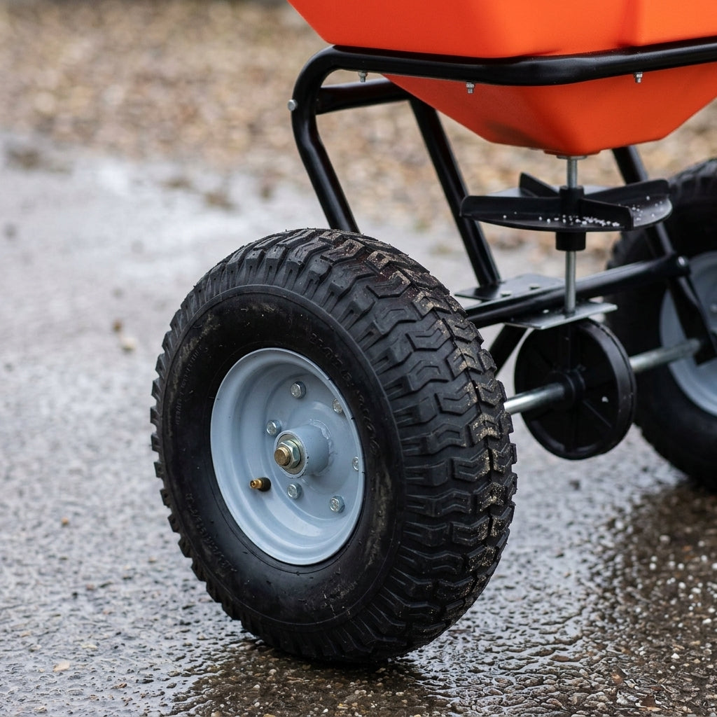 Close-up of the knobbly pneumatic tyre, steel hub and spinner disc beneath the orange hopper of a salt spreader