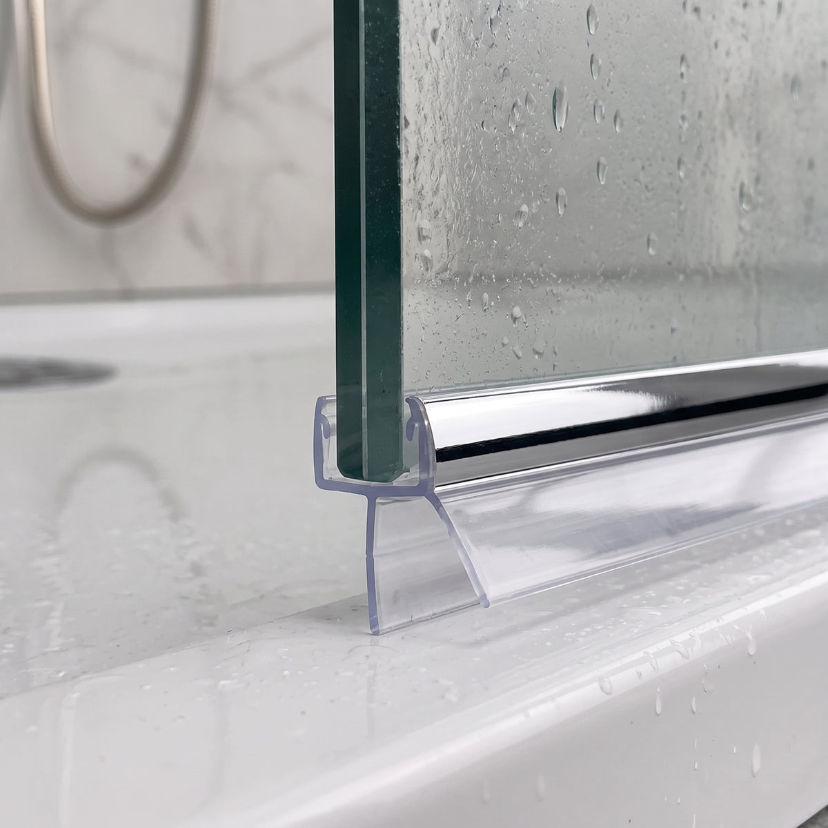 Chrome-strip seal under a wet glass screen edge on a white shower tray, marble tiles and shower hose behind