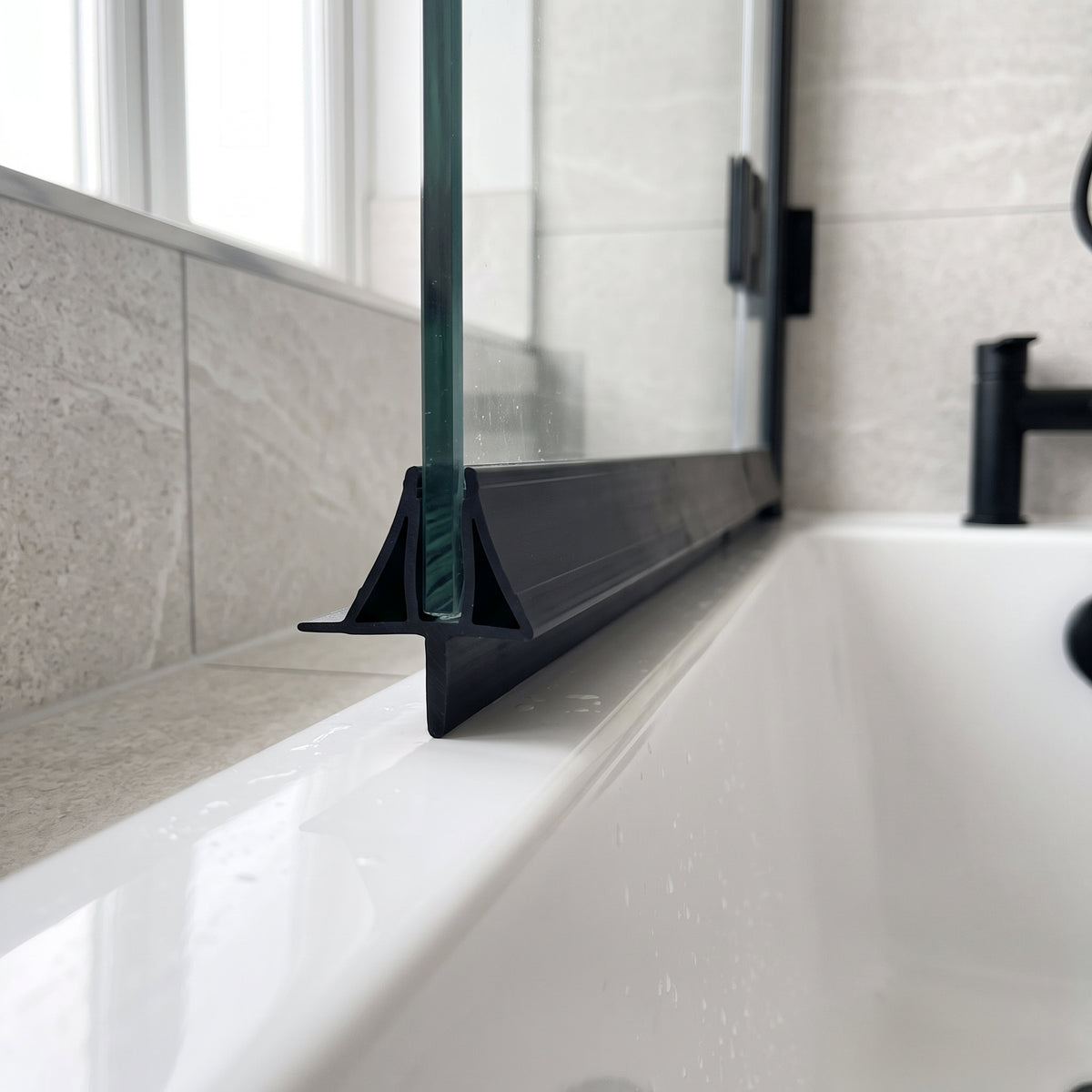 Black seal with a triangular deflector along the foot of a glass bath screen, black tap behind
