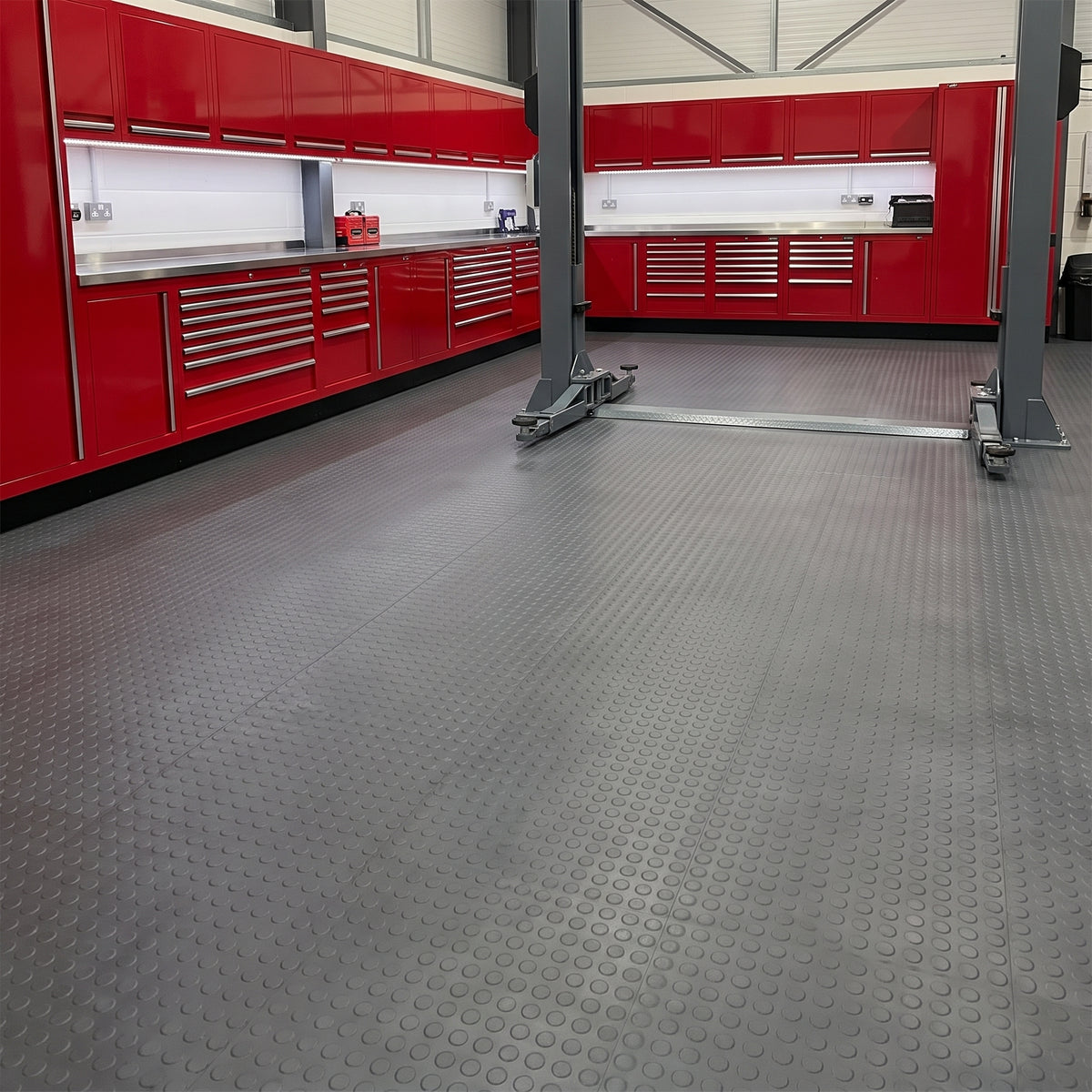 Dark grey small-dot vinyl matting laid wall to wall in continuous roll lengths across a premium workshop with red roll-cab tool chests, stainless worktop and a two-post lift