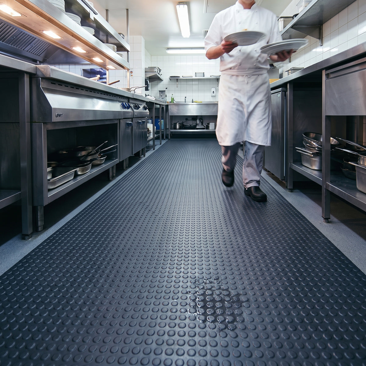 Dark grey penny-stud PVC matting runner down a restaurant kitchen walkway, a small spill held among the studs as a chef carries plates