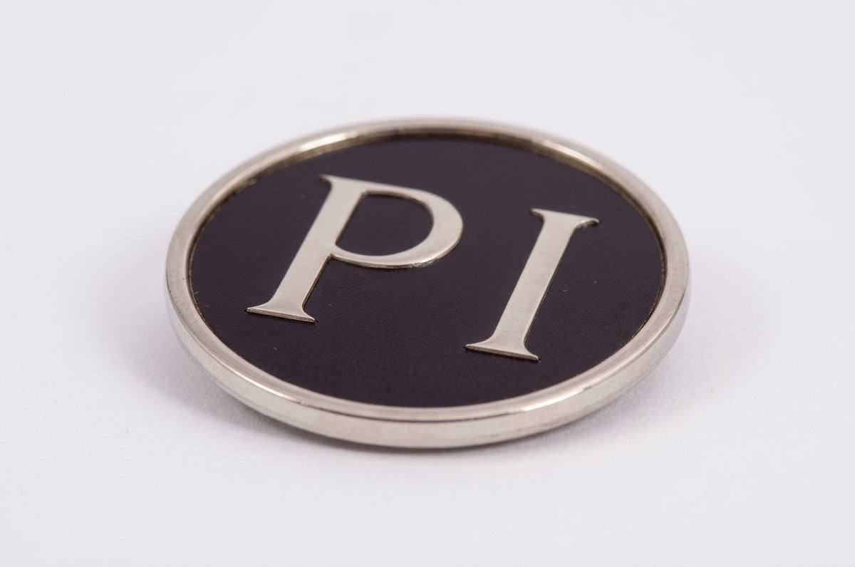 Round chrome-rimmed black badge reading PI, studio shot on white background