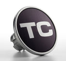 Round chrome-rimmed badge reading TC, angled studio shot on white background
