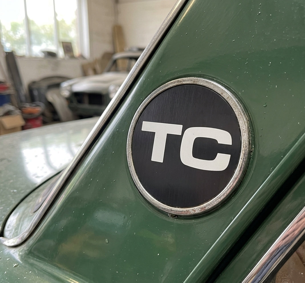Round chrome-edged TC badge fitted to the rear pillar of a green classic car