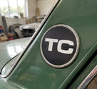 Round chrome-edged TC badge fitted to the rear pillar of a green classic car