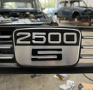 2500 S badge fitted in a slatted black grille, classic cars visible behind