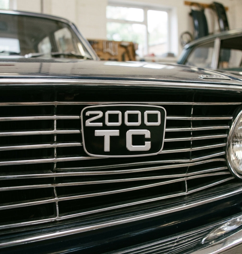 2000 TC badge in a chrome-slatted grille of a dark classic saloon, headlamp and workshop window behind
