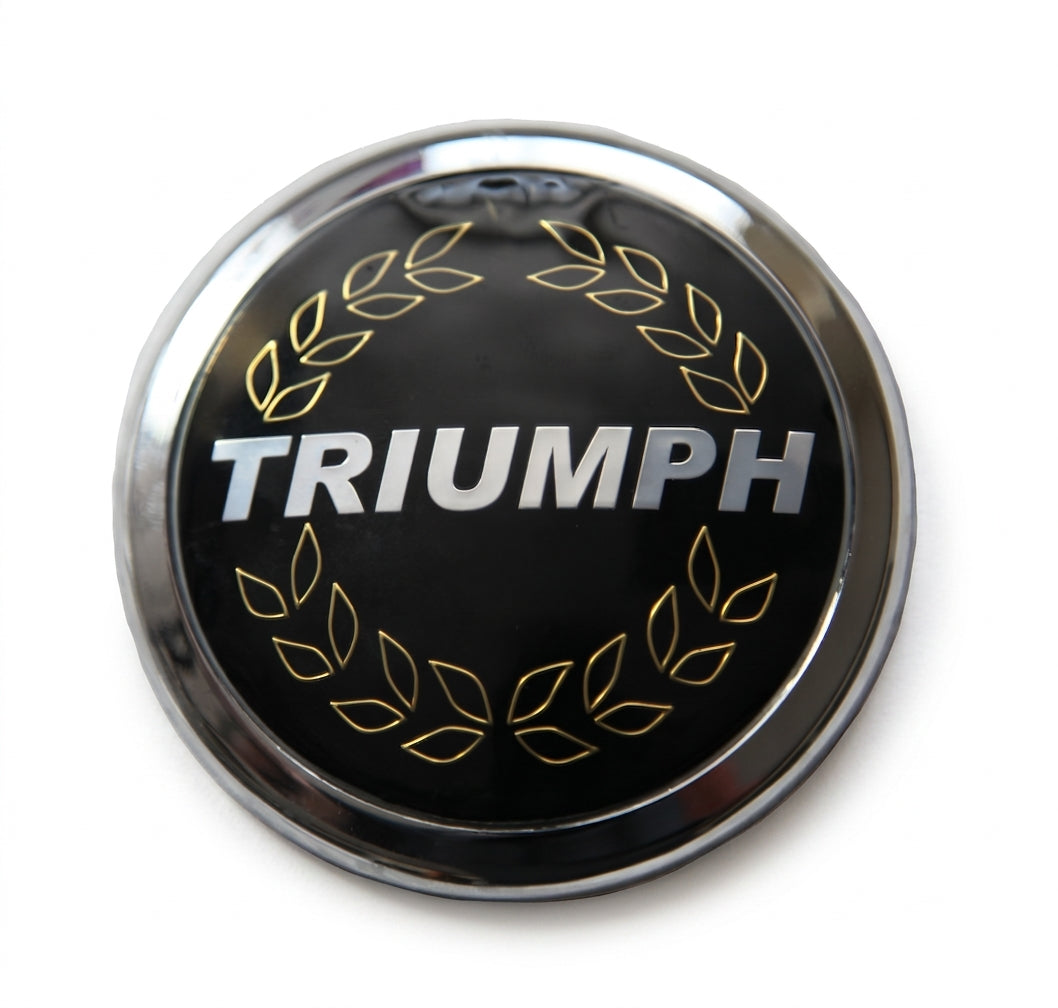 Round chrome laurel wreath badge with TRIUMPH lettering, studio shot on white