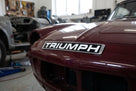 Chrome TRIUMPH script badge fitted to a maroon car's bonnet in a workshop