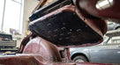 Underside of a maroon seat base tipped up, black perforated diaphragm hooked into the frame, workshop behind