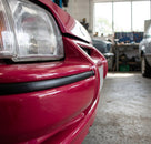 Satin black trim fitted along a red car's bumper edge, close-up workshop shot