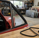 Black quarterlight seal fitted around a red Escort's rear door window, on a workbench