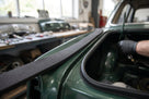 Hand applying black sponge strip to the boot lid edge of a green classic car in a workshop