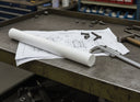 White UHMWPE rod on engineering drawings on a steel bench beside callipers, a hammer and bolts