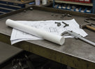 White UHMWPE rod on engineering drawings on a steel bench beside callipers, a hammer and bolts