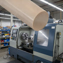 Beige PEEK rod composited large above a blue and grey CNC lathe in a bright workshop