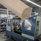 Beige PEEK rod composited large above a blue and grey CNC lathe in a bright workshop