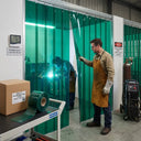 Welder in a leather apron at a green PVC strip curtain bay, arc flashing behind and a roll on a trolley