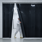 Worker in whites and a hard hat stepping through a wide black strip curtain in a white-tiled hall