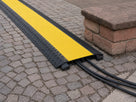 Black cable protector with closed yellow chequer lid on block paving, black cables running out from under the end