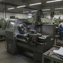 Grey CNC turning lathe on a workshop floor with an operator at a bench behind