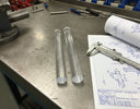 Two clear acrylic rods on a steel bench beside a graph-paper drawing, callipers and a vice