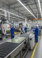 Pale green laminate panels on a roller conveyor in a machine shop, staff in blue overalls, robot arm left