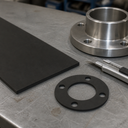 Rectangle of black rubber sheet, round gasket and knife on a steel workbench beside a machined flange