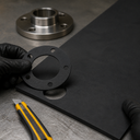 Gloved hand lifting a bolt-hole gasket from a black rubber sheet on a grey bench, flange and snap-off knife behind