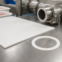 White silicone sheet and a ring gasket cut from it on a stainless bench, food plant pipework behind