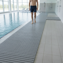 Grey PVC duckboard runner along an indoor pool edge, bather walking barefoot towards the shower area