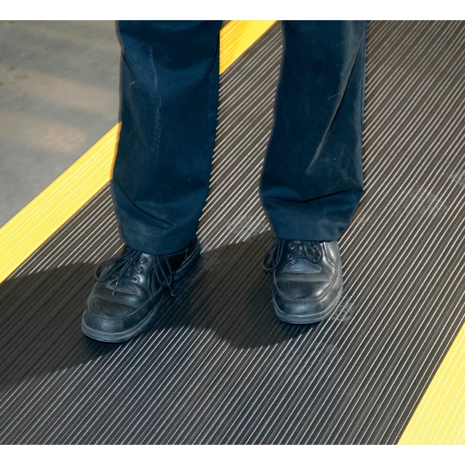 Close-up of a worker standing on a black anti-fatigue mat with yellow safety borders, highlighting the ribbed non-slip surface. The durable PVC foam cushioned mat provides anti-fatigue support for standing workstations or garages.