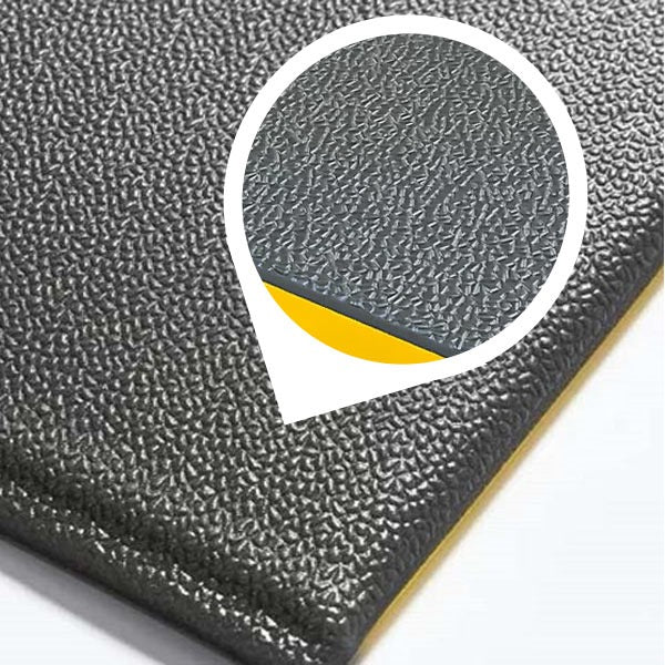 Close-up shot of the corner of a heavy-duty anti-fatigue mat. The surface features a dark grey, pebbled, non-slip texture. An inset bubble magnifies the thickness and shows the wear-resistant PVC foam construction with a visible yellow bottom layer, ideal for cushioned support in workstations and industrial garages.
